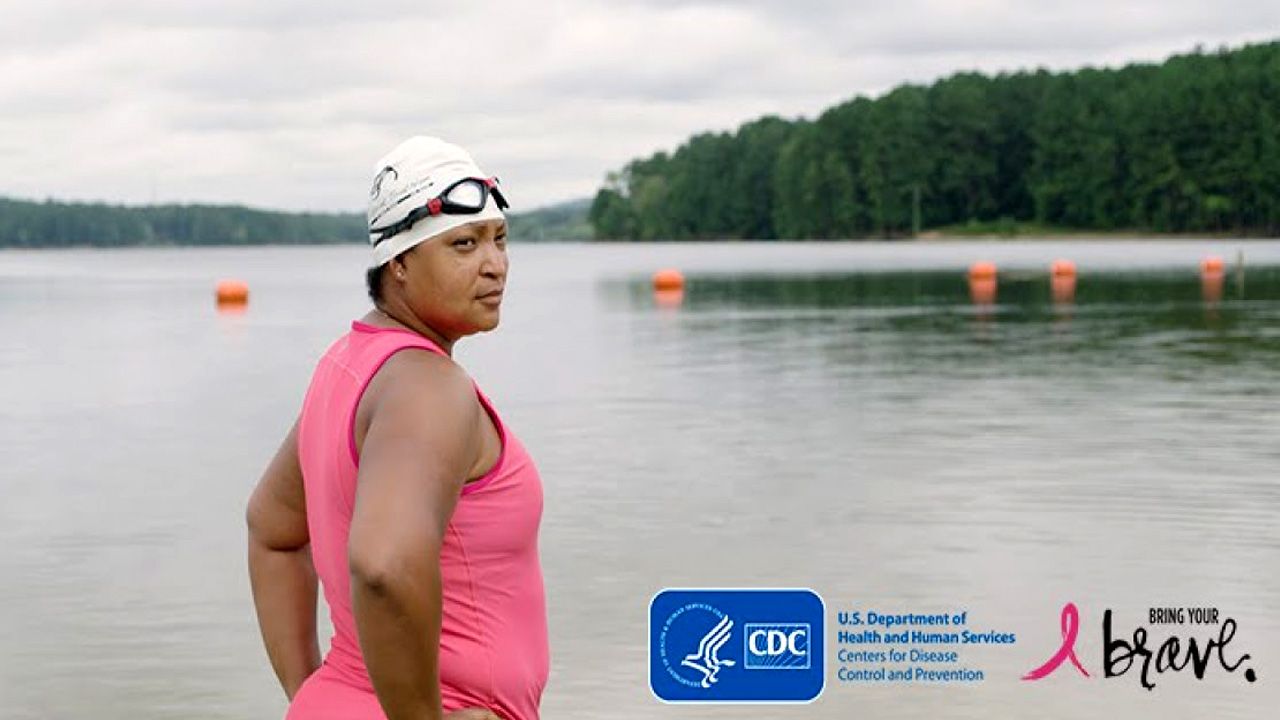 A breast cancer survivor in a bathing suit and swim cap stands in front of a lake and looks back with confidence.